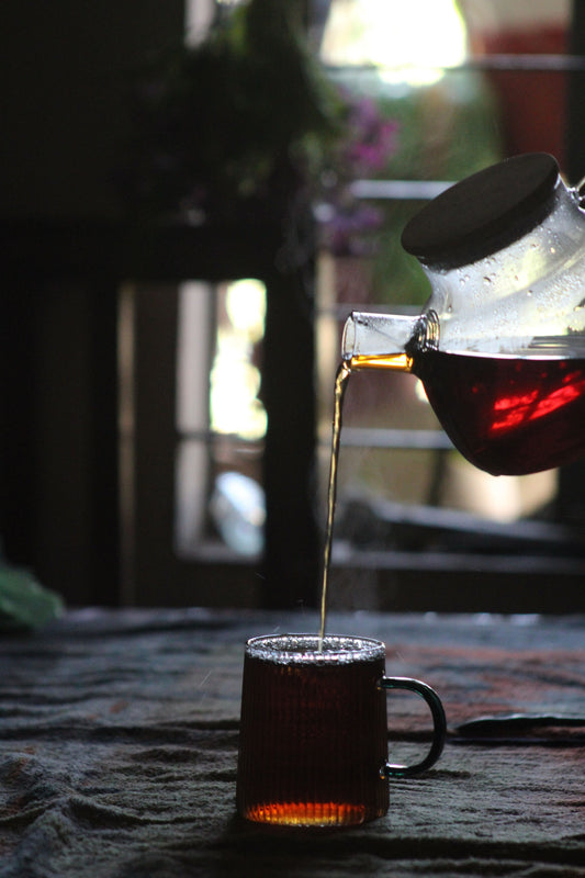 Tea being poured from a glass teapot into a glass mug, warm amber liquid catching evening light, moody dark background