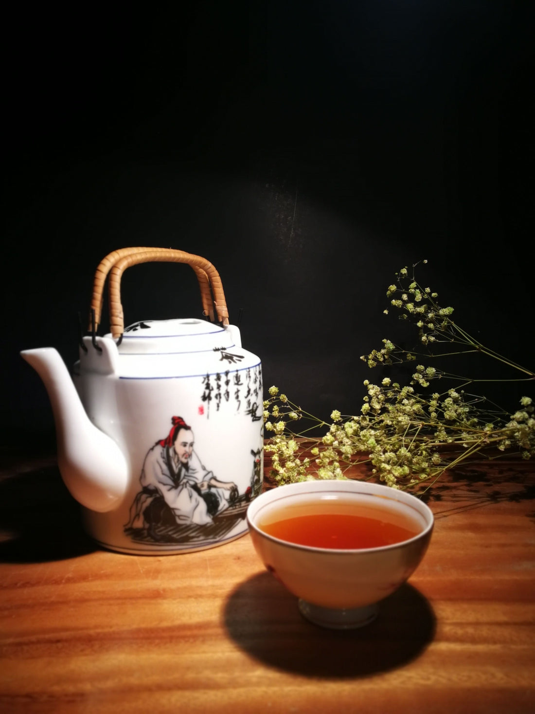 Traditional Chinese ceramic teapot with painted figure and calligraphy, small cup of brewed tea beside it, dried flowers in the background, warm wood surface against a dark backdrop