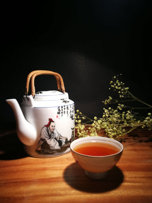Traditional Chinese ceramic teapot with painted figure and calligraphy, small cup of brewed tea beside it, dried flowers in the background, warm wood surface against a dark backdrop